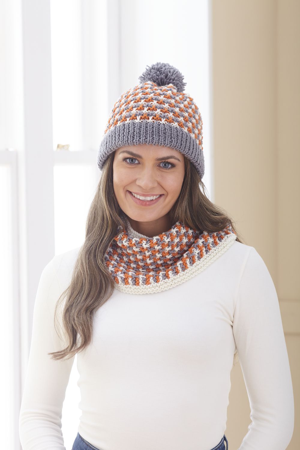 A woman is wearing a knitted hat and cowl in grey, cream and orange.