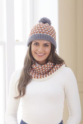 A woman is wearing a knitted hat and cowl in grey, cream and orange.