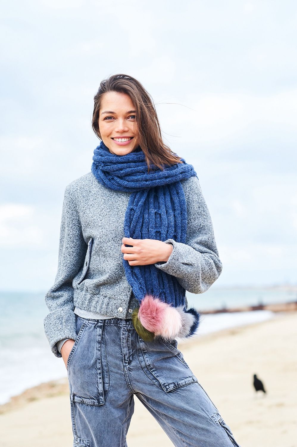 A woman is wearing a dark blue knitted scarf with faux fur pompoms on one end.