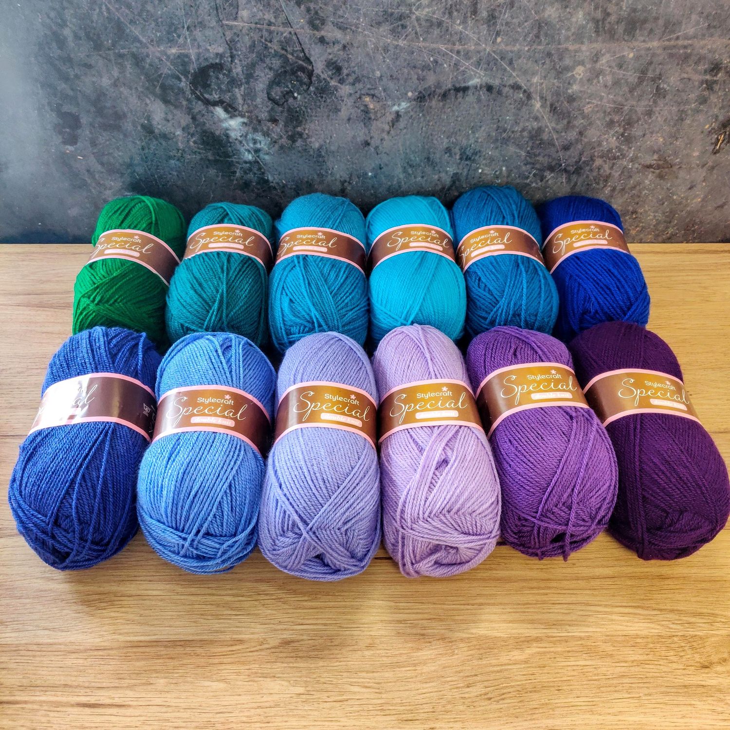 12 balls of yarn in shades of green, blue, teal and purple laid out on a wooden surface.