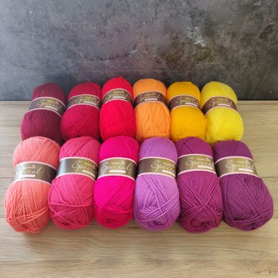 12 balls of yarn in shades of red, orange, yellow, pink and purple laid out on a wooden surface.