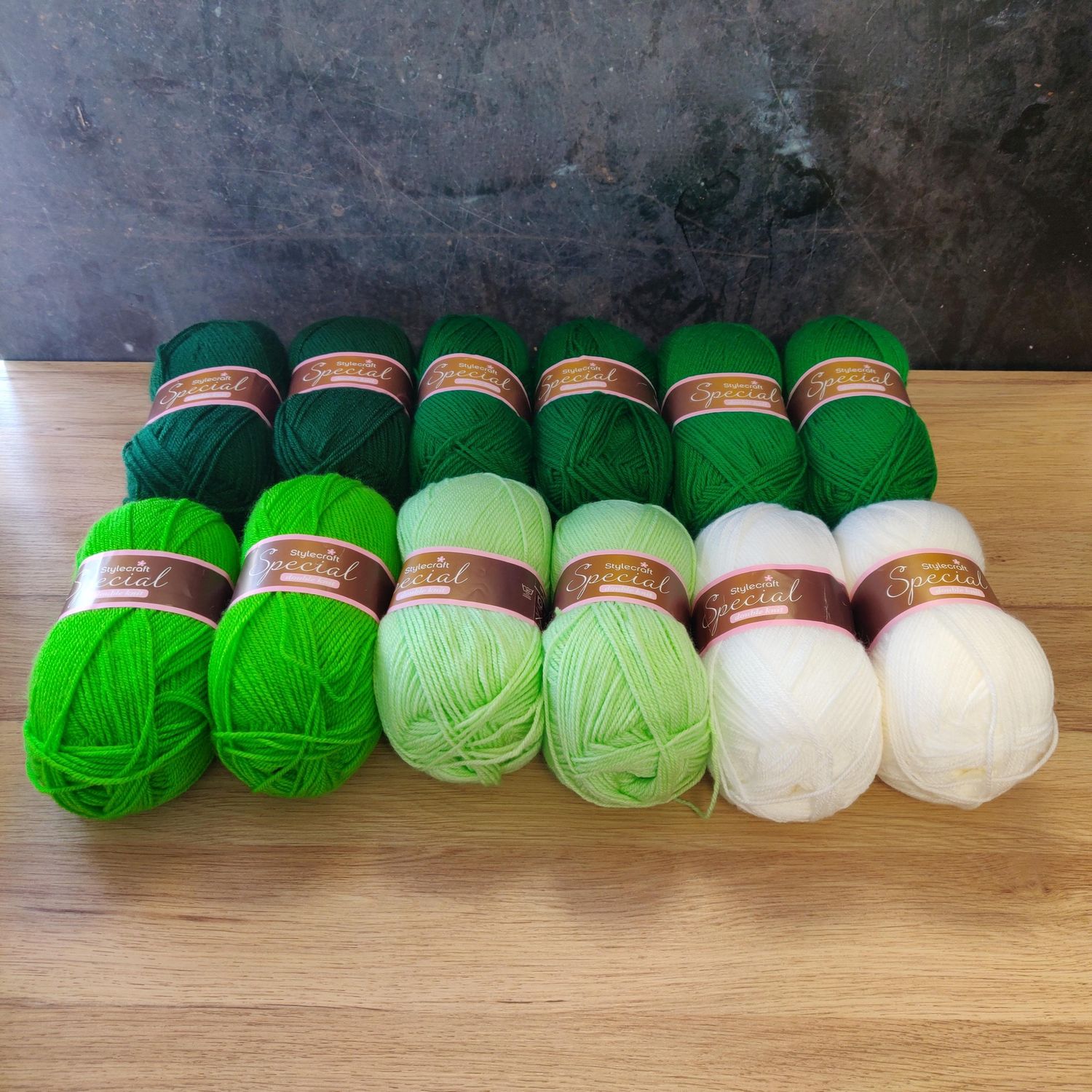 12 balls of yarn in shades of green and white laid out on a wooden surface.