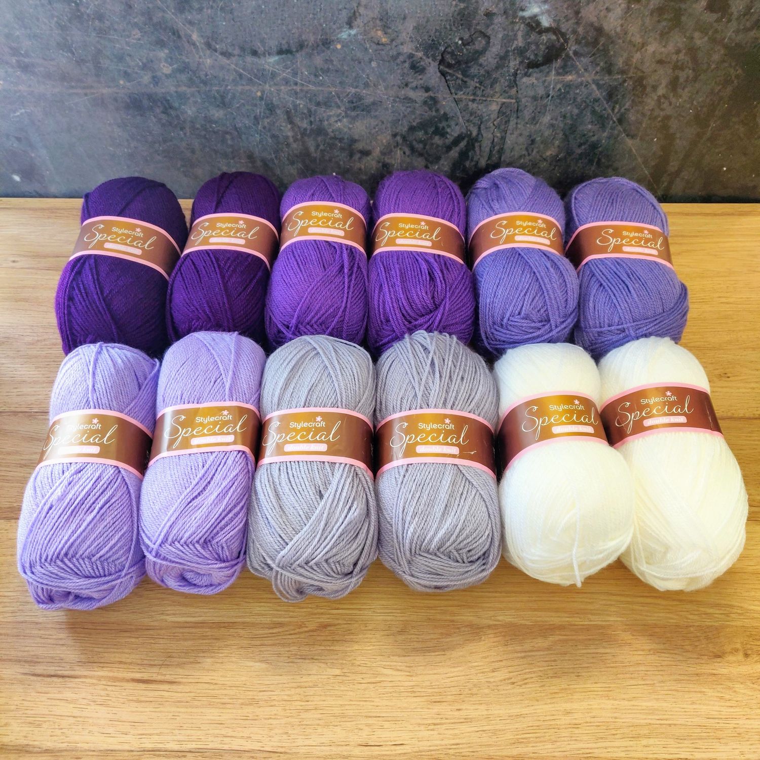 12 balls of yarn in shades of purple and white laid out on a wooden surface.