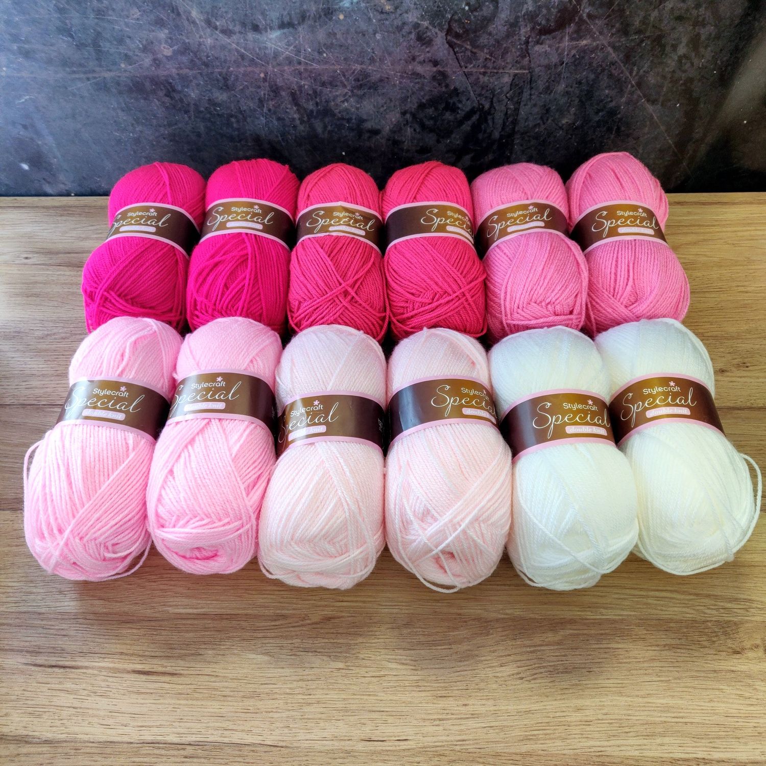 12 balls of yarn in shades of pink and white laid out on a wooden surface.