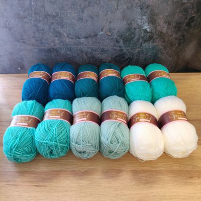 12 balls of yarn in shades of teal, aqua and white laid out on a wooden surface.