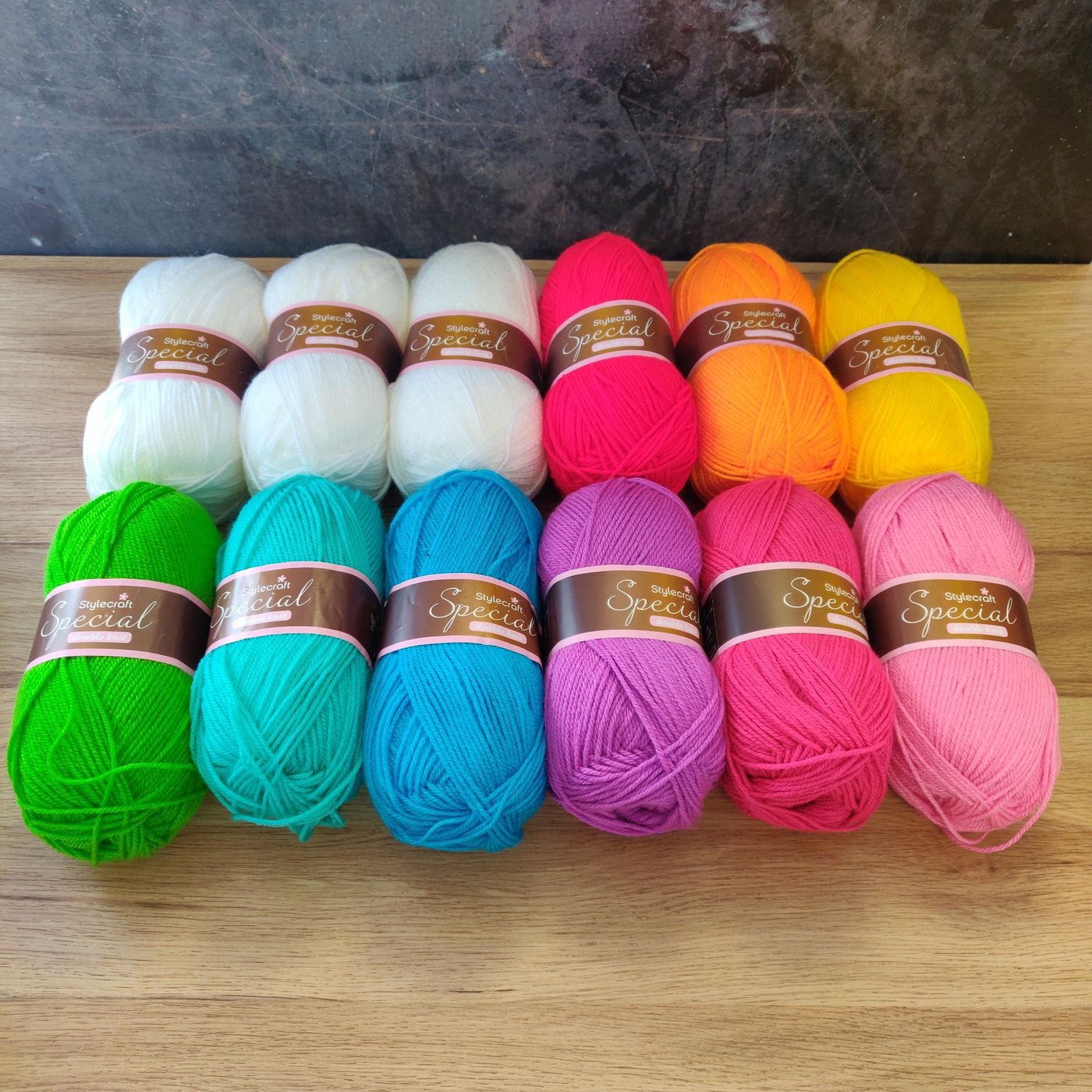 12 balls of yarn in shades of white, red, orange, yellow, green, blue, purple and pink laid out on a wooden surface.