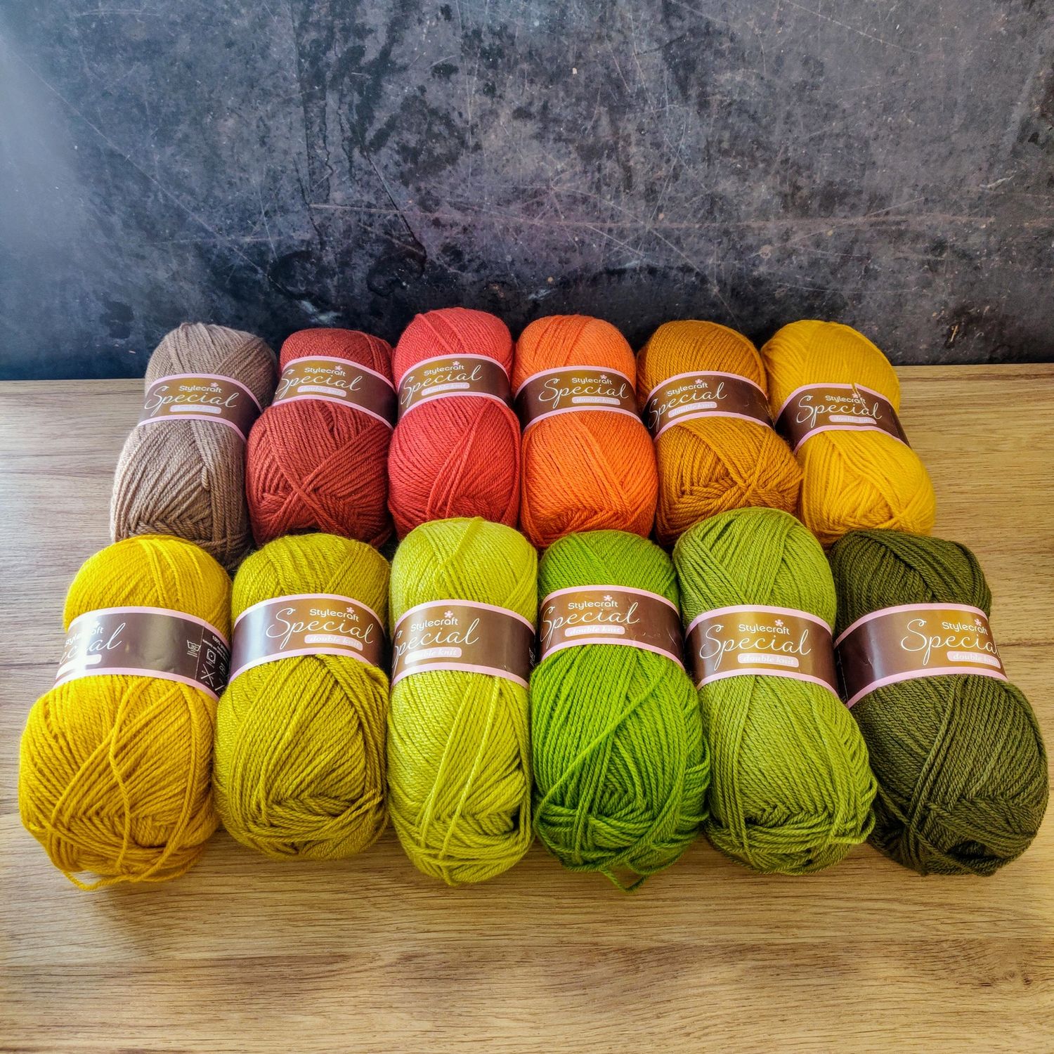 12 balls of yarn in shades of brown, red, orange, mustad and green laid out on a wooden surface.