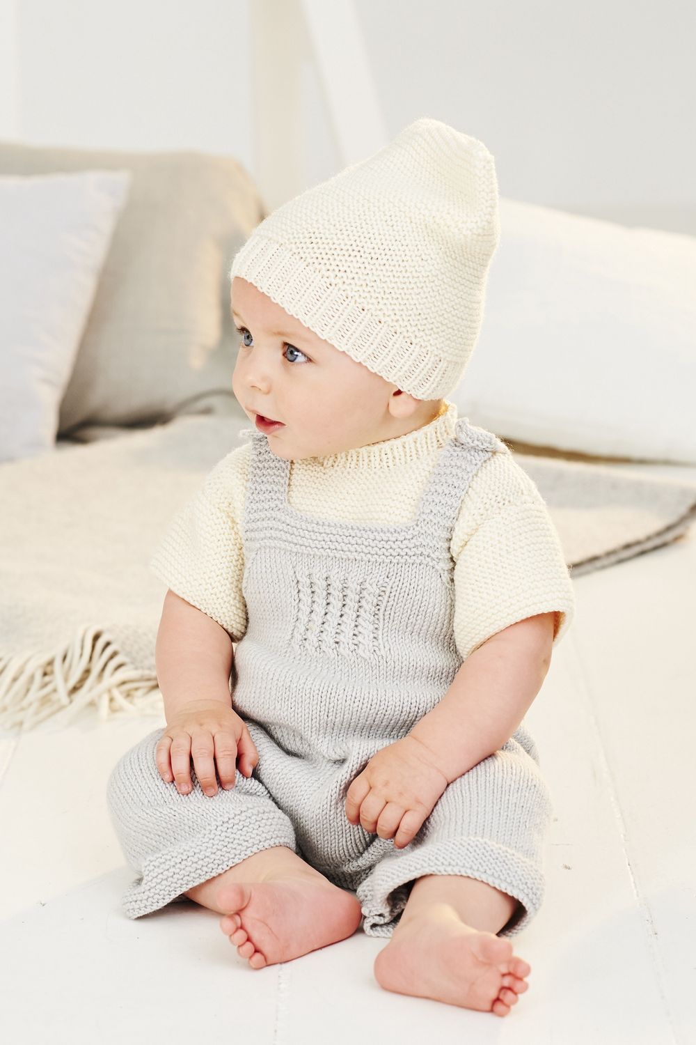 A baby is wearing a cream knitted t-shirt and hat and light grey knitted dungarees.