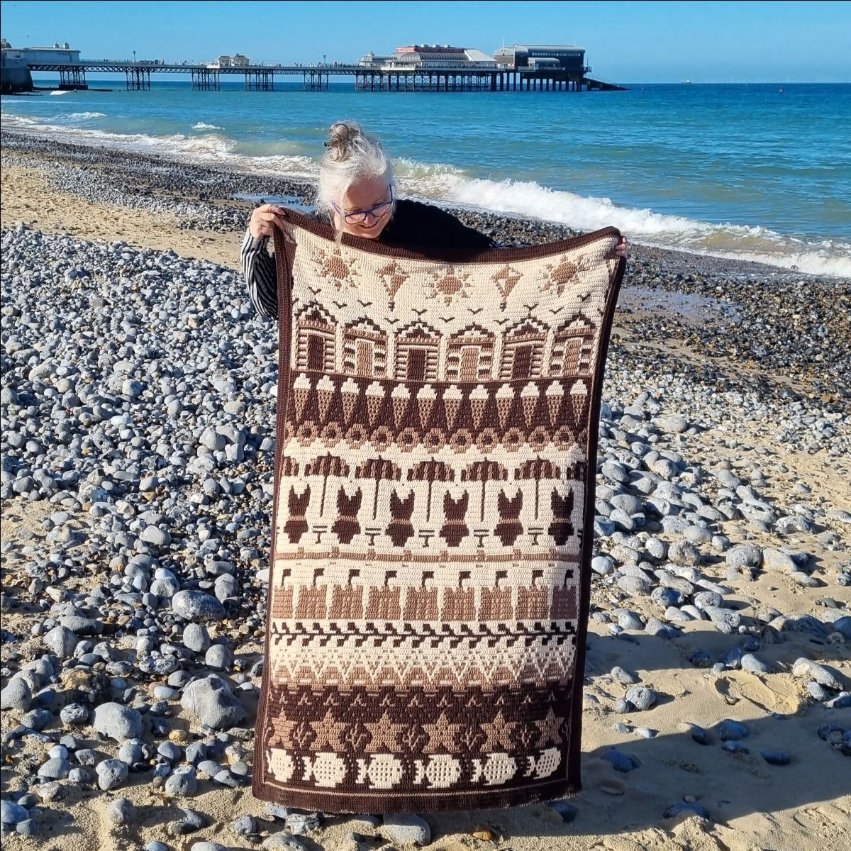 A crocheted blanket in cream and brown. The blanket contains various seaside designs such as beach huts, ice creams, sand castles, starfish and more.