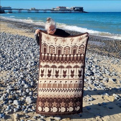 A crocheted blanket in cream and brown. The blanket contains various seaside designs such as beach huts, ice creams, sand castles, starfish and more.