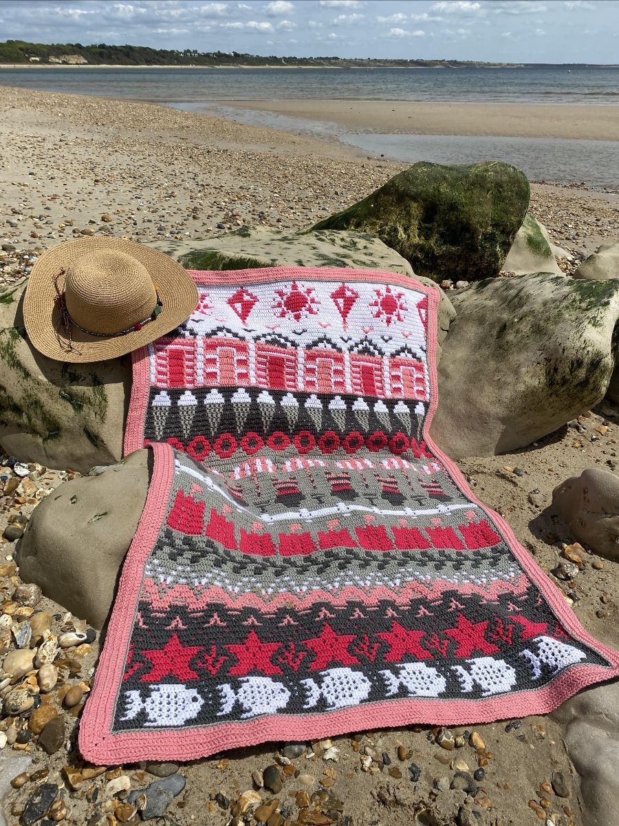 A crocheted blanket in white, pink and grey. The blanket contains various seaside designs such as beach huts, ice creams, sand castles, starfish and more.