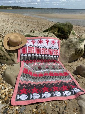 A crocheted blanket in white, pink and grey. The blanket contains various seaside designs such as beach huts, ice creams, sand castles, starfish and more.