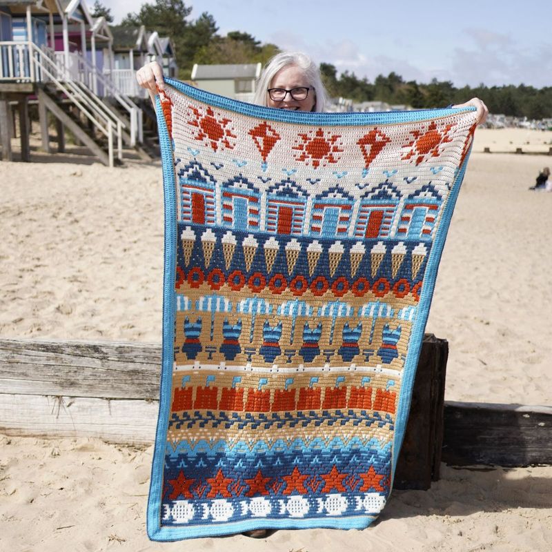 A crocheted blanket in cream, blue, brown and red. The blanket contains various seaside designs such as beach huts, ice creams, sand castles, starfish and more.