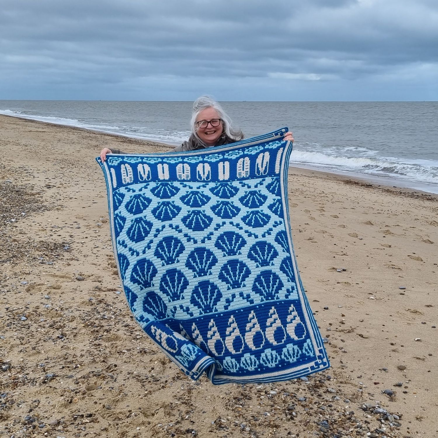 A woman standing on a beach holding the blanket, made in various shades of blue.
