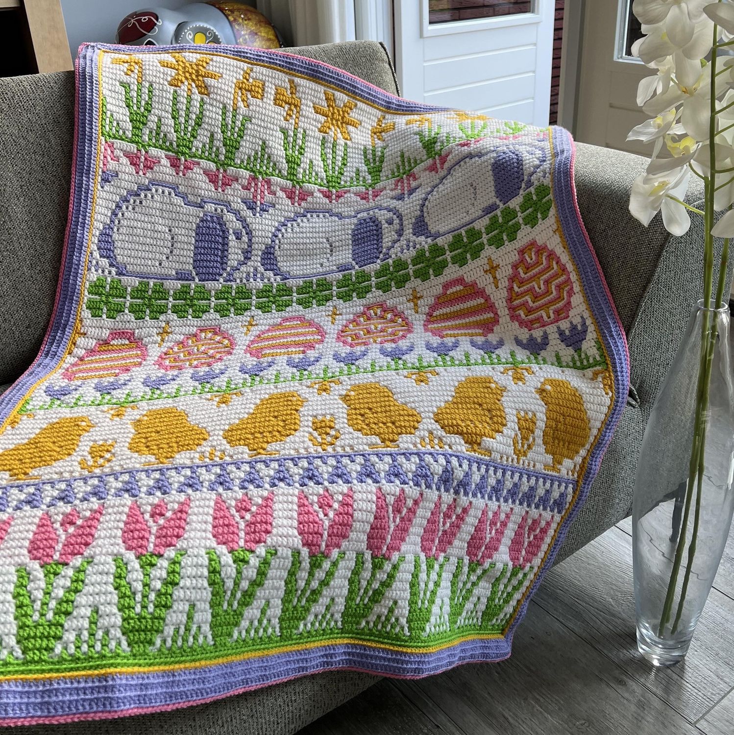 A crocheted blanket in white, green, pink, purple and yellow draped over a sofa. The blanket contains various Easter designs such as rabbits, eggs, chicks, tulips and more.