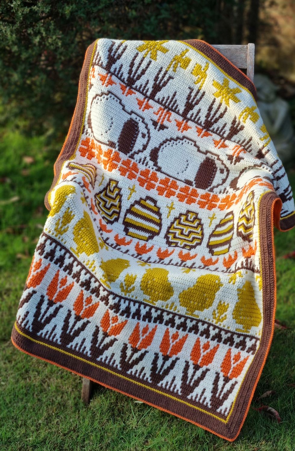 A crocheted blanket in cream, mustard, orange and brown. The blanket contains various Easter designs such as rabbits, eggs, chicks, tulips and more.