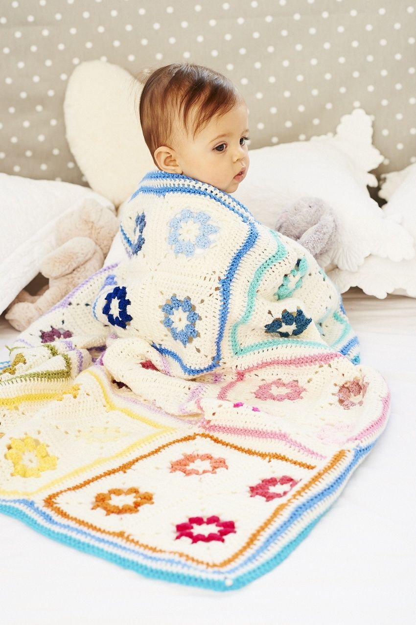 A baby is wearing a crocheted blanket, made up of white squares with colourful flower motifs in the centre of each.