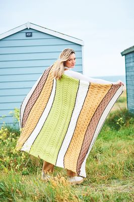 A woman is holding a cable knit blanket in shades of green, cream, brown and yellow.