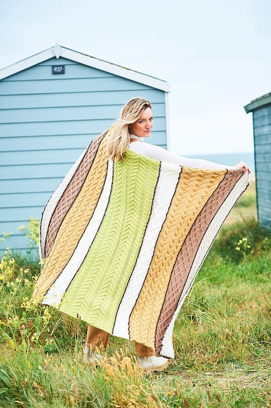 A woman is holding a cable knit blanket in shades of green, cream, brown and yellow.