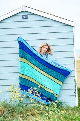 A woman is holding a cable knit blanket in shades of green, teal and blue.