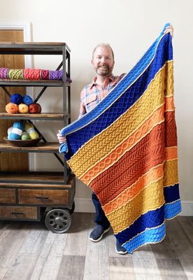 A man is holding a cable knit blanket in shades of blue, orange and yellow.
