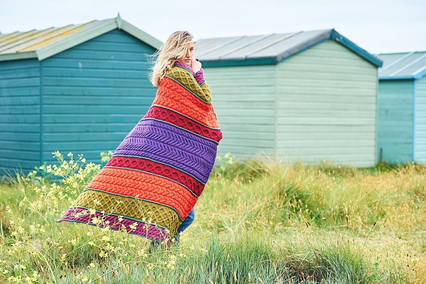 A woman is holding a cable knit blanket in shades of green, red, orange and purple.