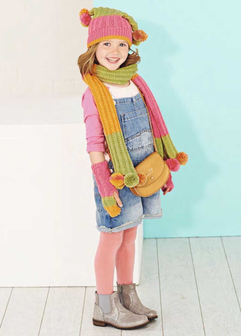 A girl is wearing a knitted hat, scarf and mittens in a variegated pink, orange and green yarn.