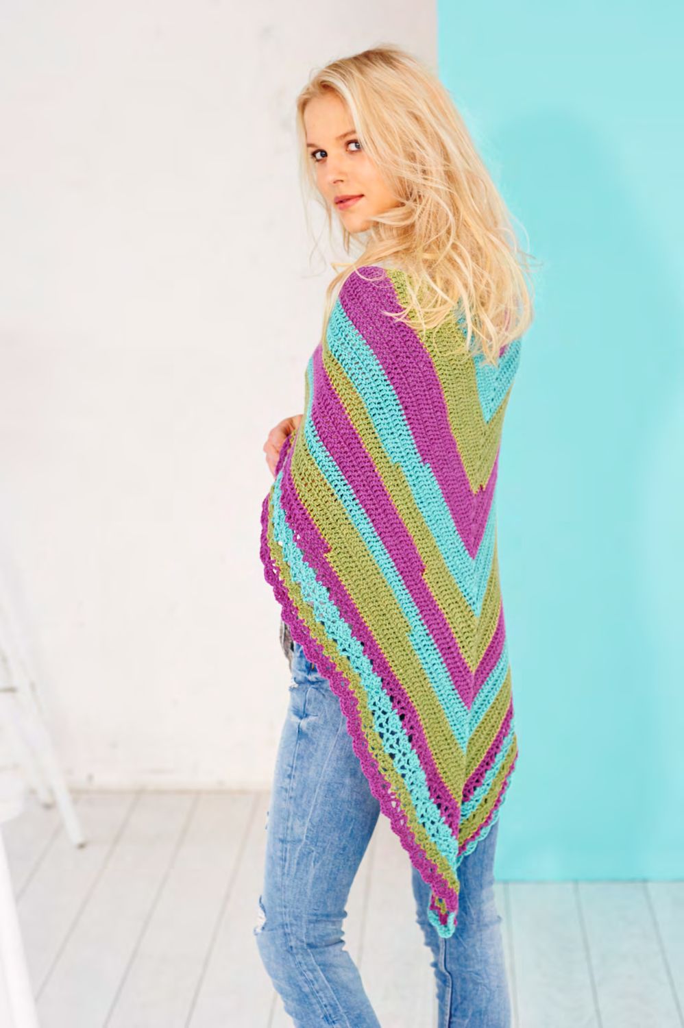 A woman is wearing a crocheted shawl in a variegated purple, blue and green yarn.