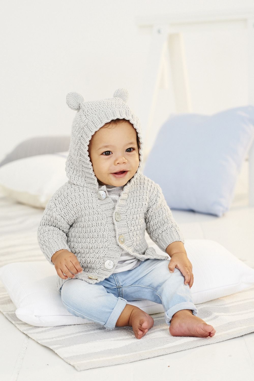 A baby is wearing a light grey knitted hooded cardigan, with bear ears on the hood.