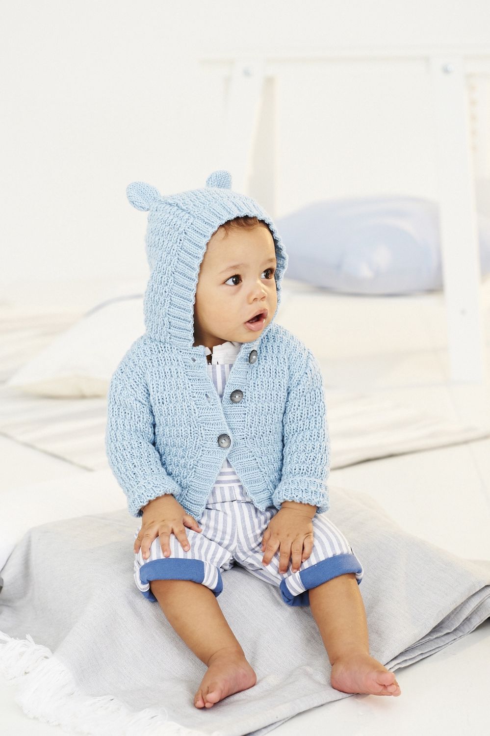 A baby is wearing a light blue knitted hooded cardigan, with bear ears on the hood.