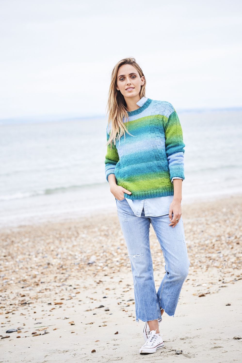A woman is wearing a knitted sweater in a variegated green and blue yarn.