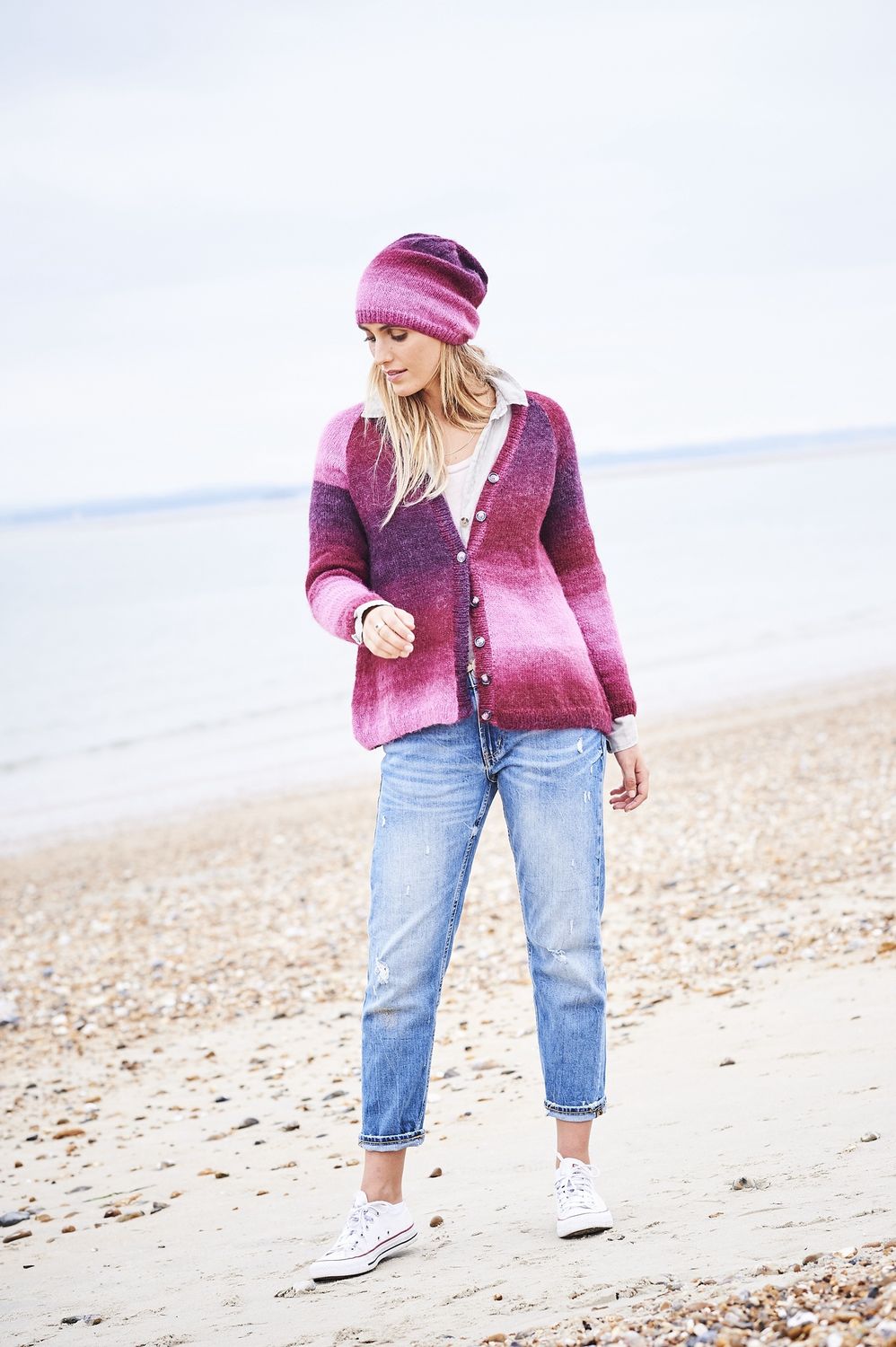 A woman is wearing a knitted cardigan and matching hat in a variegated pink yarn.