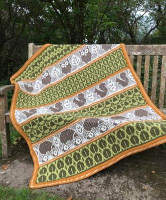 A crocheted blanket featuring autumnal patterns such as owls, squirrels, acorns and more in shades of cream, green and brown.