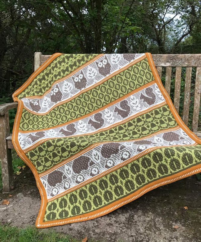 A crocheted blanket featuring autumnal patterns such as owls, squirrels, acorns and more in shades of cream, green and brown.