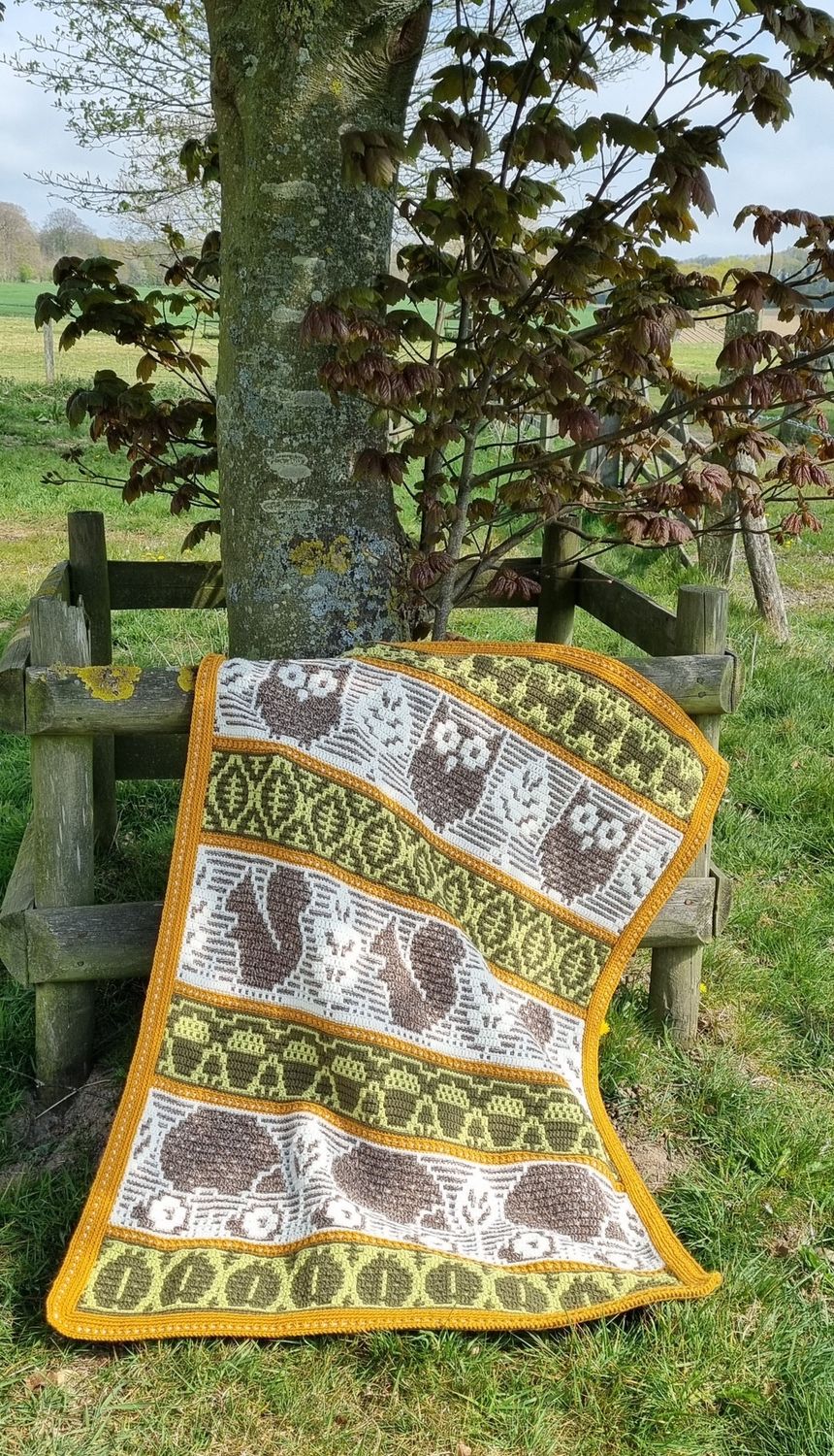 A crocheted blanket featuring autumnal patterns such as owls, squirrels, acorns and more in shades of cream, green and brown.