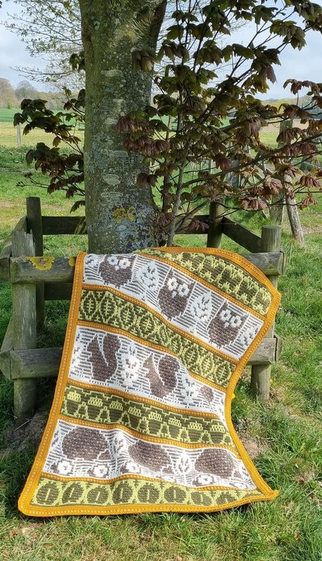 A crocheted blanket featuring autumnal patterns such as owls, squirrels, acorns and more in shades of cream, green and brown.