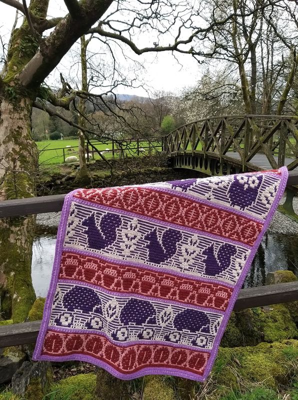 A crocheted blanket featuring autumnal patterns such as owls, squirrels, acorns and more in shades of cream, pink and purple.