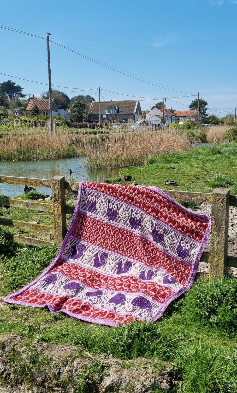 A crocheted blanket featuring autumnal patterns such as owls, squirrels, acorns and more in shades of cream, pink and purple.