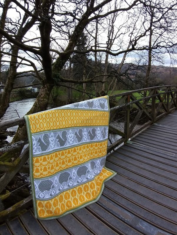 A crocheted blanket featuring autumnal patterns such as owls, squirrels, acorns and more in shades of white, yellow and grey.