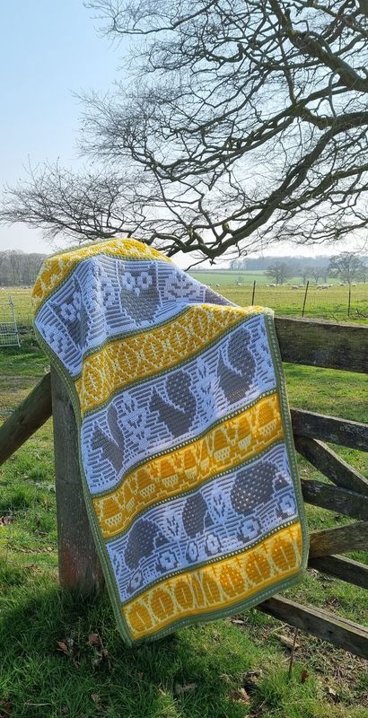 A crocheted blanket featuring autumnal patterns such as owls, squirrels, acorns and more in shades of white, yellow and grey.