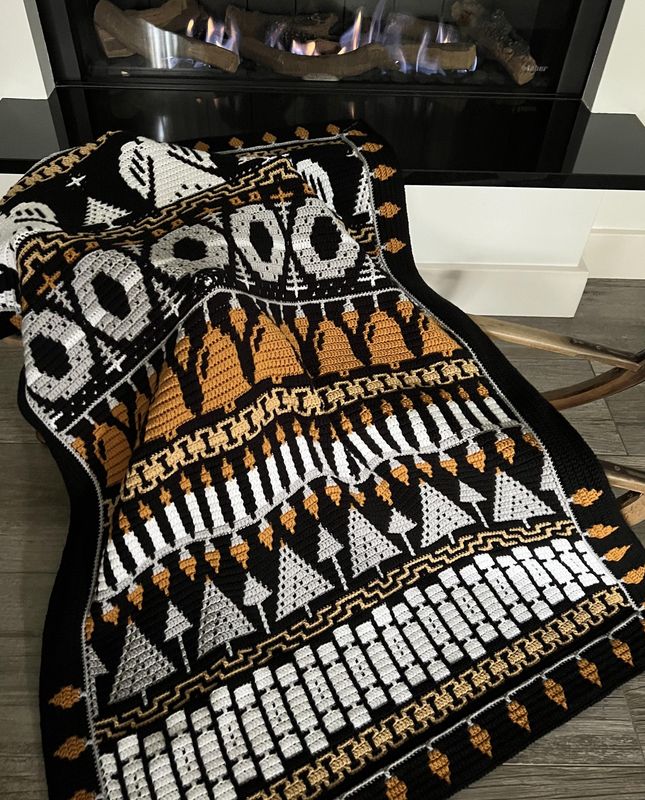 A crocheted blanket in black, white, grey and brown. The blanket contains various Christmas designs such as angels, wreaths, candles, crackers and more.