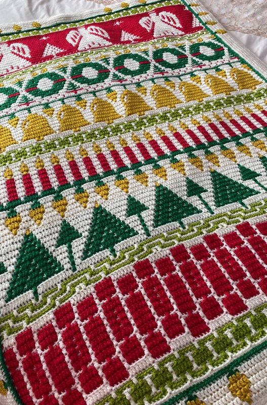 A crocheted blanket in cream, green, red and yellow. The blanket contains various Christmas designs such as angels, wreaths, candles, crackers and more.