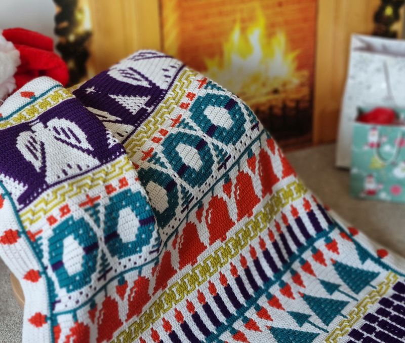 A crocheted blanket in silver, purple, green, blue and red. The blanket contains various Christmas designs such as angels, wreaths, candles, crackers and more.