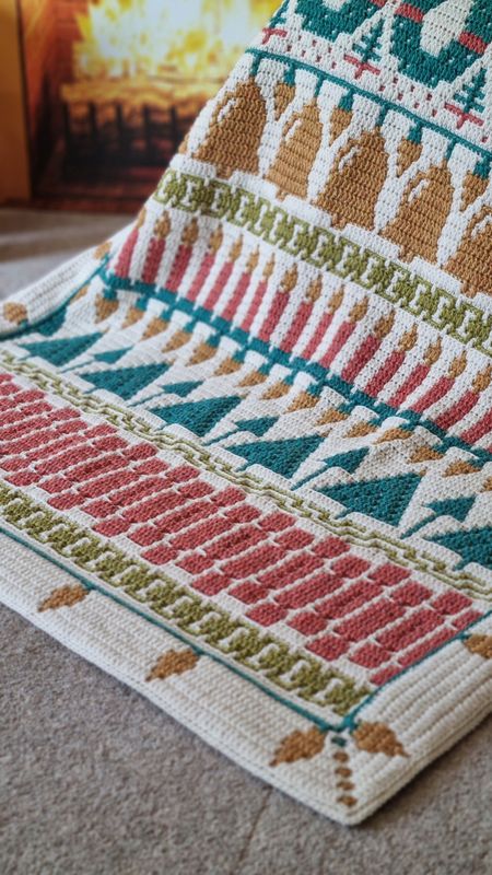 A crocheted blanket in cream, green, yellow, teal and red. The blanket contains various Christmas designs such as angels, wreaths, candles, crackers and more.