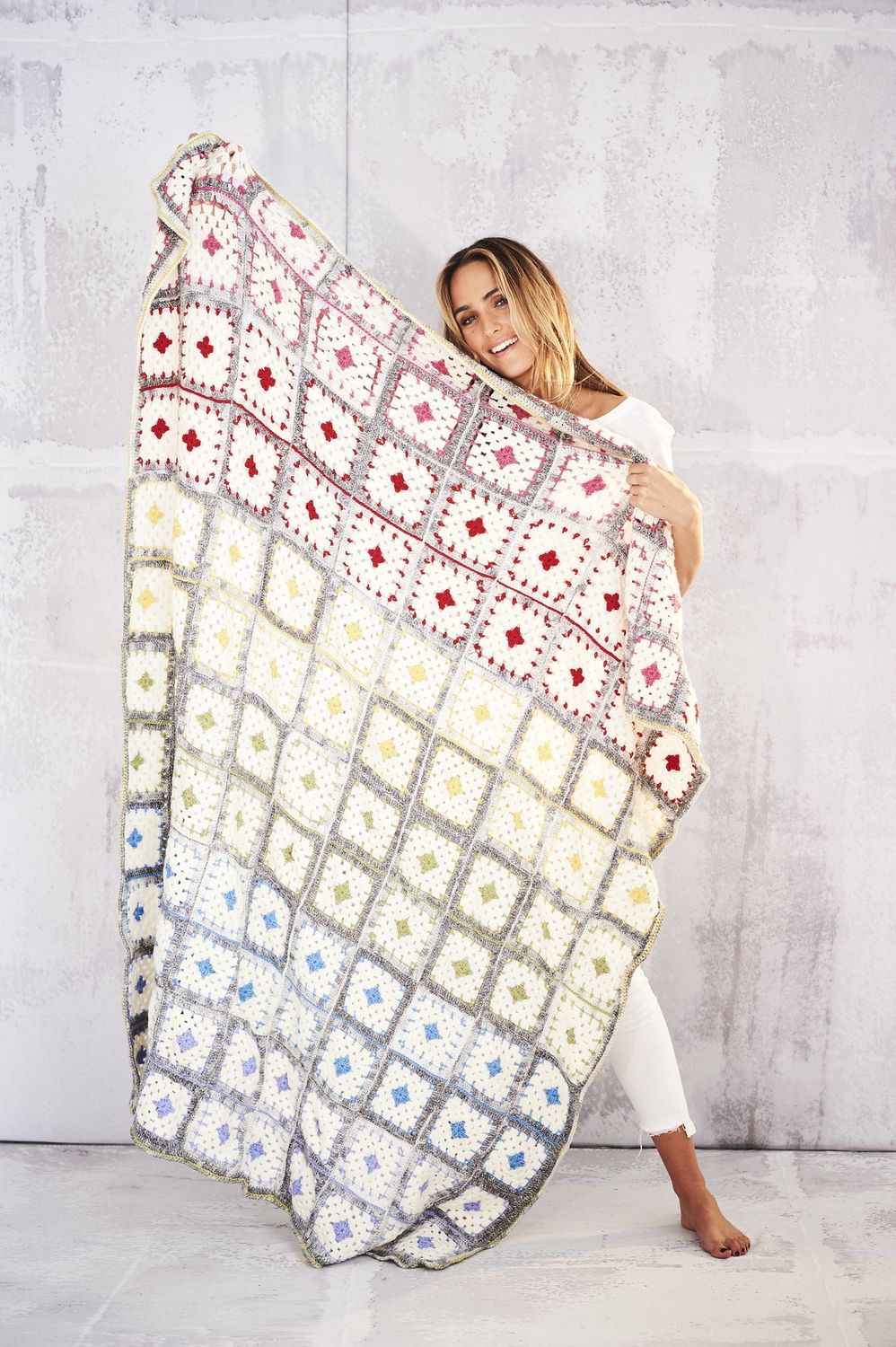 A woman is holding a crocheted blanket made up of cream and grey squares with brightly coloured centres.