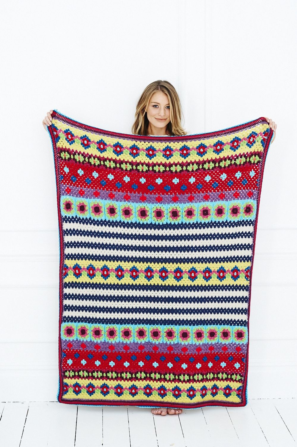 A woman is holding a crocheted blanket in blue, red, yellow, purple, green and cream.