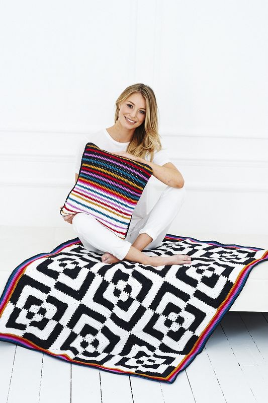 A woman is holding a black, white, red, purple, gold and teal crocheted cushion while sitting on a crocheted blanket made up of black and white crocheted squares.
