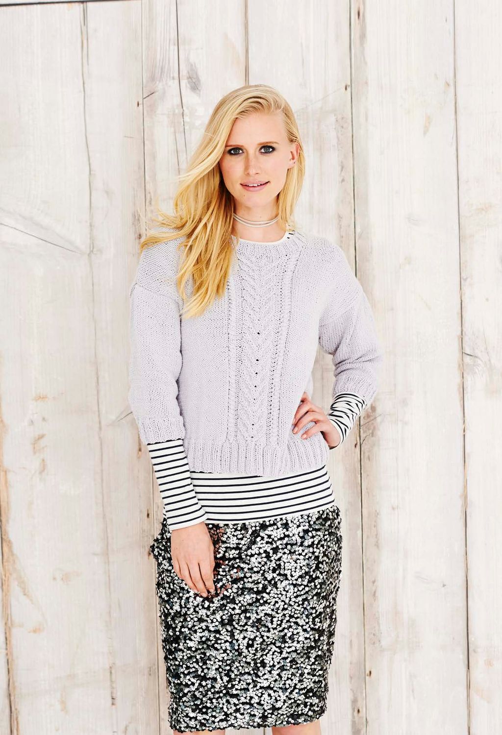 A woman is wearing a light grey knitted sweater with cable down the front centre.