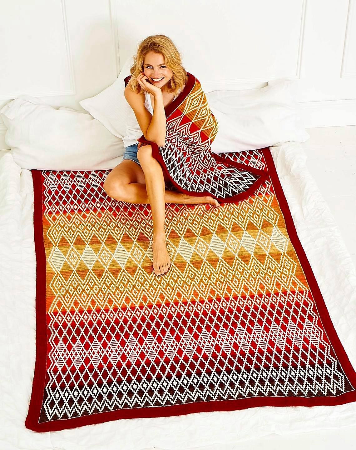 A woman is sitting on the large version of this blanket in reds, yellows and cream, while the small version in the same colourway is draped around her shoulders.
