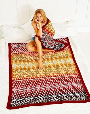 A woman is sitting on the large version of this blanket in reds, yellows and cream, while the small version in the same colourway is draped around her shoulders.